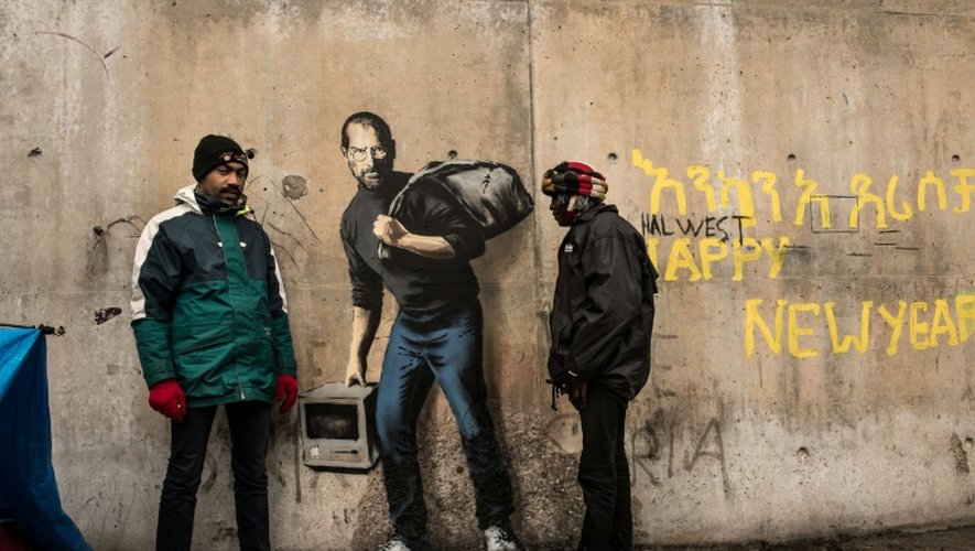 Des migrants devant une oeuvre de l'artiste britannique Banksy photographiée le 12 décembre 2015 et située à l'entrée de la "Jungle" de Calais, représente Steve Jobs (Apple), qui porte un baluchon et un vieil ordinateur