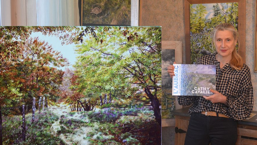 Cathy présentant son livre "Les couleurs de l’Aubrac à la pointe de mon pinceau"à côté de l’un de ses nombreux tableaux.